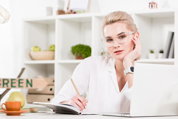 donna in occhiali che scrive in notebook vicino a computer portatile in ufficio, concetto di risparmio ambientale
  - Foto, immagini
