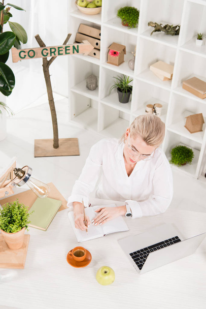 overhead view of woman writing in notebook near laptop with go green sign behind, environmental saving concept  - Photo, Image