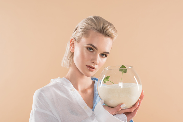 beautiful girl holding fish bowl with sand and small green plant isolated on beige, environmental saving concept  - Photo, Image