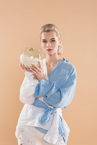 beautiful woman holding fish bowl with sand and small green plant while standing in eco clothing isolated on beige, environmental saving concept  - Photo, Image