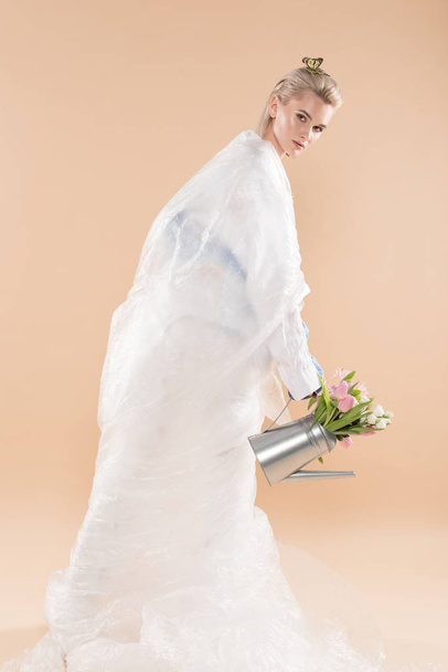 attractive toung woman wrapped in polyethylene holding watering can with flowers isolated on beige, environmental saving concept  - Foto, afbeelding
