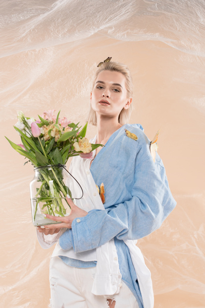 attractive girl standing with butterflies on eco clothing and holding glass vase with flowers on beige background, environmental saving concept  - Photo, Image