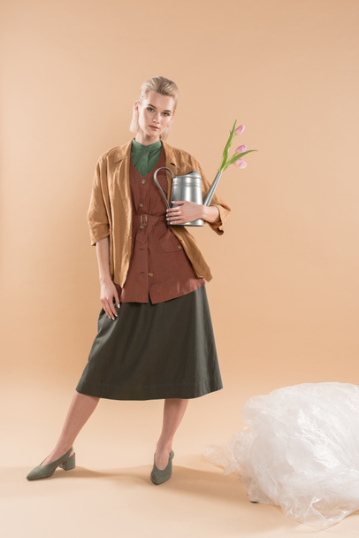attractive girl in eco clothing holding watering can with flowers near polyethylene on beige background, environmental saving concept - Valokuva, kuva