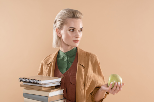 attractive girl holding books and green apple while standing in eco clothing isolated on beige - Fotó, kép