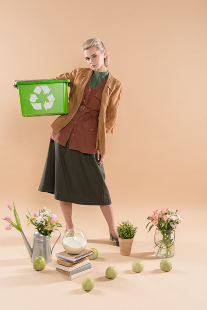bella donna che tiene scatola di riciclaggio vicino a piante e fiori su sfondo beige, concetto di risparmio ambientale
 - Foto, immagini