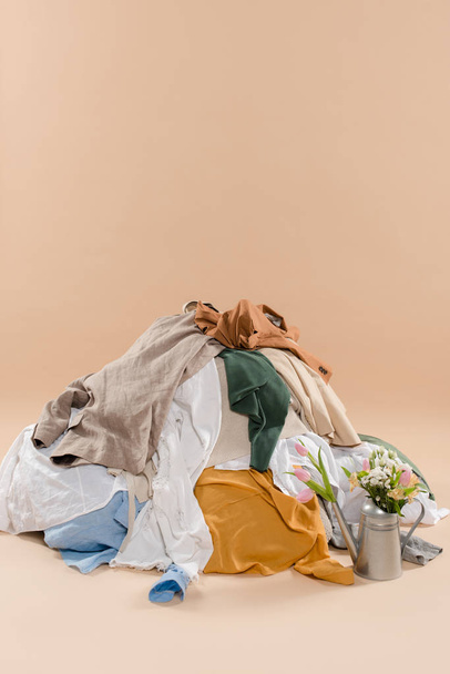 stack of clothing near watering can with flowers on beige background, environmental saving concept - Foto, imagen