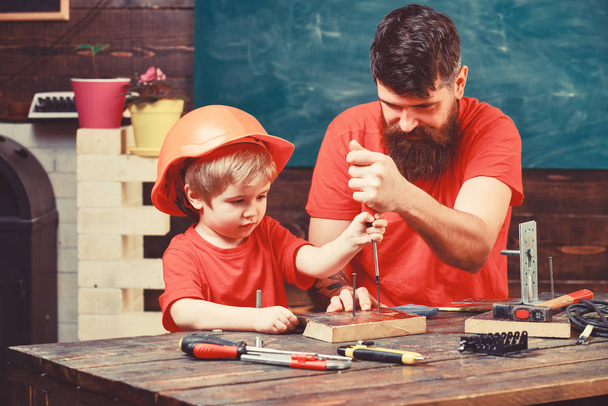 Teamwork in workshop concept. Father, parent with beard teaching little son to use tool screwdriver. Boy, child busy in protective helmet learning to use screwdriver with dad - Foto, Imagem