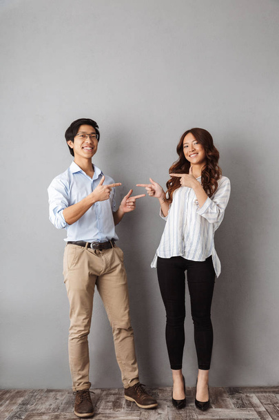 Full length of cheerful asian couple standing over gray background, pointing fingers at copy space - Photo, Image