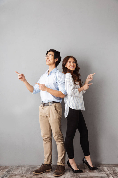 Piena lunghezza di allegra coppia asiatica in piedi su sfondo grigio, puntando il dito contro spazio copia
 - Foto, immagini