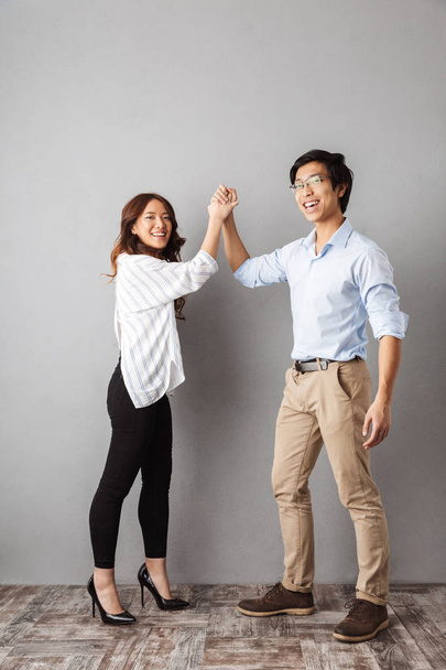 Full length of cheerful asian couple standing over gray background, giving high five - Foto, afbeelding