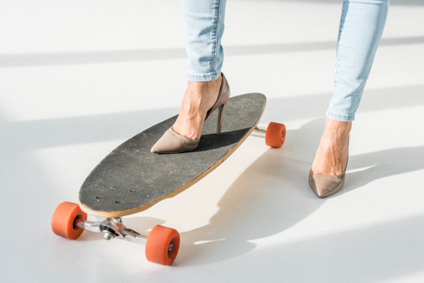 cropped view of woman in high heels putting leg on skateboard on grey background - 写真・画像