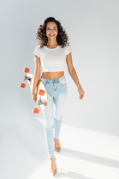 cheerful woman with curly hair holding skateboard while walking on grey background - Foto, imagen