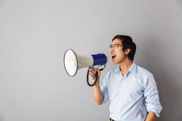 Angry asian business man standing isolated over gray background, screaming at a loudspeaker - Foto, Bild