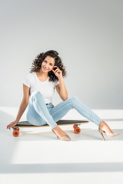 attractive girl with curly hair sitting on skateboard in high heels on grey background - 写真・画像