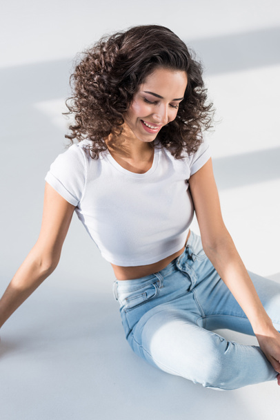 Pretty curly girl in jeans sitting on floor with smile - Fotografie, Obrázek