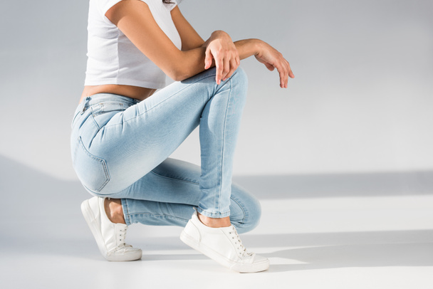 Cropped view of stylish woman in jeans sitting on grey background - Zdjęcie, obraz