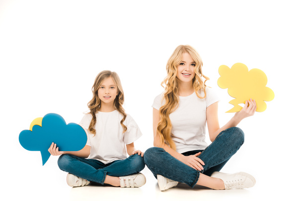 Lächelnde Mutter und Tochter sitzen mit überkreuzten Beinen auf dem Boden und halten Sprechblasen auf weißem Hintergrund - Foto, Bild