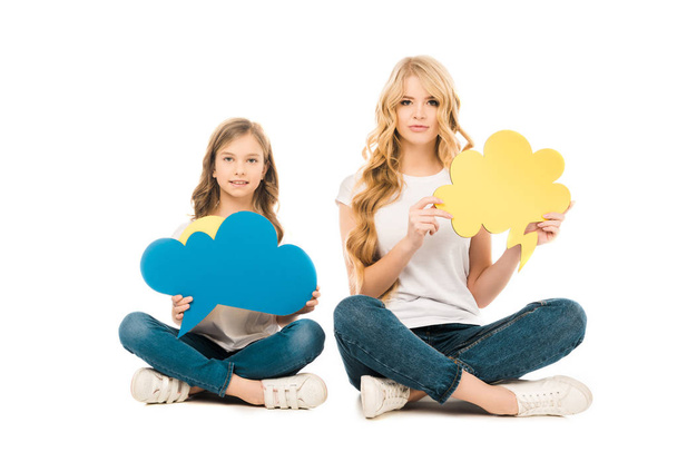 pretty mother and cute daughter sitting on floor with crossed legs and holding thought bubbles while looking at camera on white background - Photo, Image