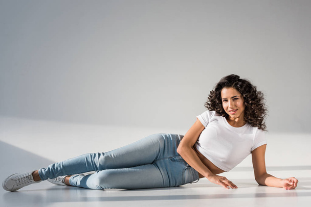 Beautiful curly girl in jeans lying on grey background - 写真・画像