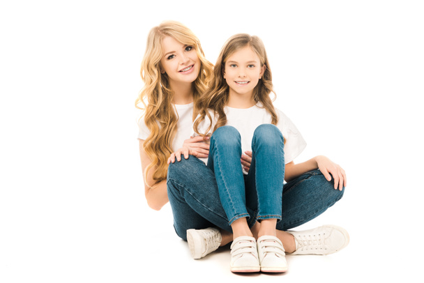 smiling mother and daughter sitting on floor and looking at camera on white background - Foto, Imagen