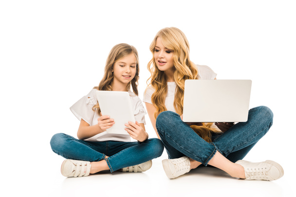 Uśmiechnięte matki i córki za pomocą laptopa i tabletu cyfrowy, siedząc na podłodze ze skrzyżowanymi nogami na białym tle - Zdjęcie, obraz