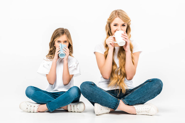 mother and daughter sitting on floor with crossed legs and drinking coffee on white background - Photo, Image