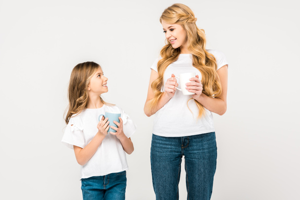 smiling mother and daughter holding cups and looking at each other on white background - Foto, Imagen