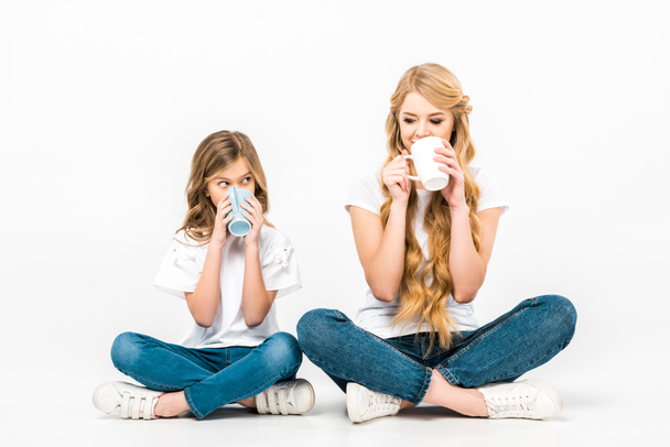 cheerful mother and daughter drinking coffee while sitting on floor with crossed legs on white background - Photo, Image