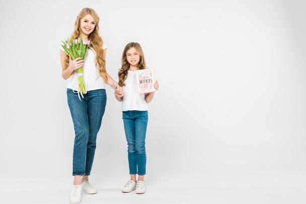 улыбающаяся мама с букетом розовых тюльпанов и улыбающаяся дочь с поздравительной открыткой на день матери держась за руки, глядя в камеру на белом фоне
 - Фото, изображение