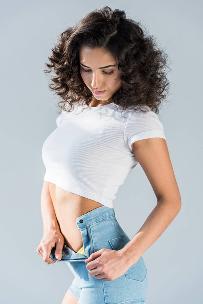 Slim curly girl unzipping shorts isolated on grey - 写真・画像