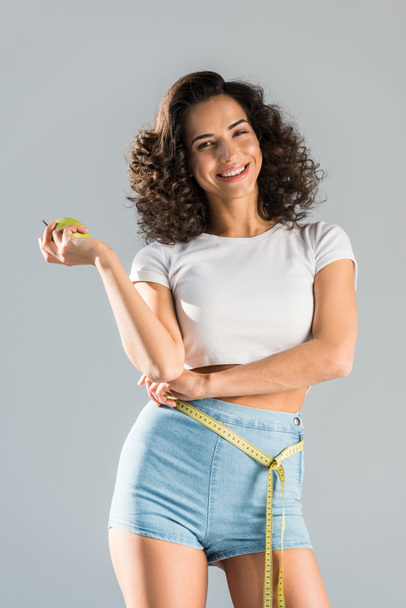 Cheerful curly girl in shorts with measuring tape holding green apple isolated on grey - Photo, Image