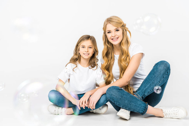 happy mother and child sitting on floor and looking at camera while soap bubbles flying around on white background - Foto, Bild