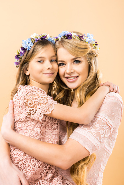 happy mother and daughter in colorful floral wreaths embracing and looking at camera on yellow background - Photo, Image