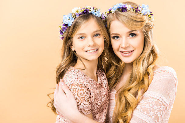 cute child and pretty mom in colorful floral wreaths embracing and looking at camera on yellow background - Photo, image