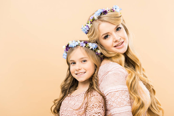 beautiful woman and cute daughter in colorful floral wreaths standing back to back and looking at camera on yellow background - Zdjęcie, obraz