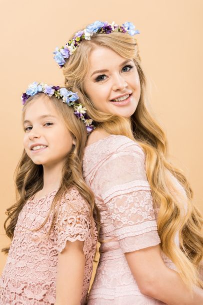 attractive mother and adorable child in colorful floral wreaths standing back to back and looking at camera on yellow background - Фото, изображение
