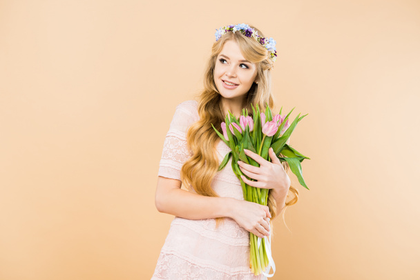 smiling woman in elegant lacy dress and floral wreath holding pink tulips and looking away on yellow background - Foto, afbeelding