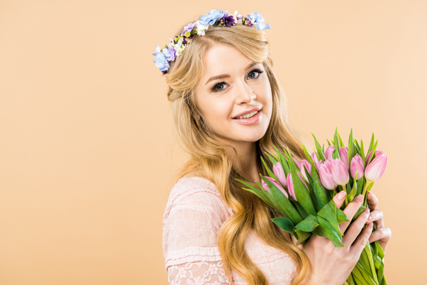 happy woman in colorful floral wreath holding pink tulips and looking at camera on yellow background - Photo, Image