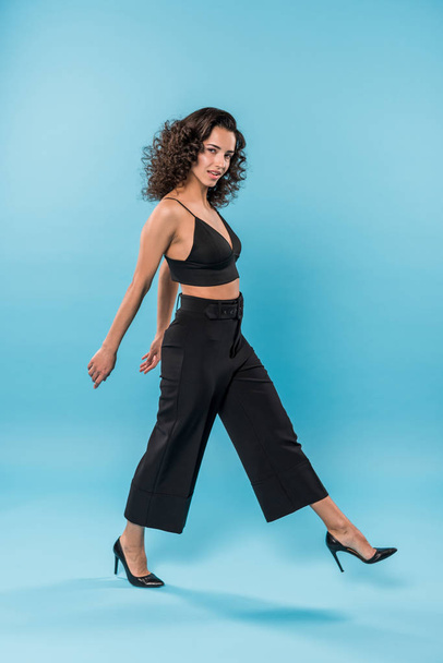 Curly young woman in black clothes walking on blue background - Photo, Image