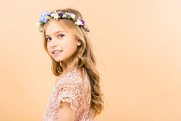 entzückendes Kind in elegantem Spitzenkleid und Blumenkranz, lächelnd und in die Kamera blickend auf gelbem Hintergrund - Foto, Bild