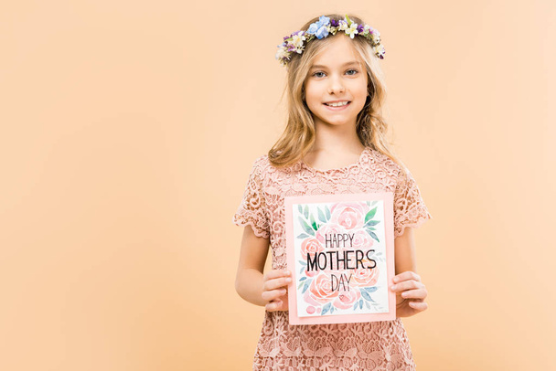 αξιολάτρευτο χαμογελώντας παιδιού σε λεπτή φόρεμα δαντελωτές και floral στεφάνι κρατώντας ευτυχισμένη μητέρες ημέρα ευχετήρια κάρτα σε κίτρινο φόντο - Φωτογραφία, εικόνα