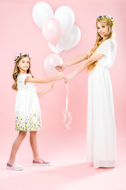 beautiful mother and cute daughter in white elegant dresses and floral wreaths holding festive air balloons on pink background - Fotografie, Obrázek