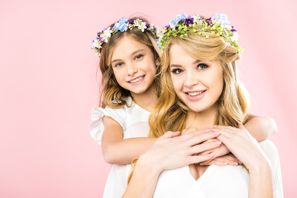 pretty woman and cute child in colorful floral wreaths smiling and looking at camera on pink background - Foto, afbeelding