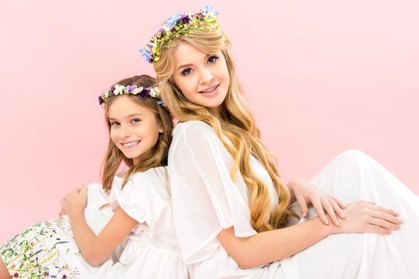 beautiful mother and adorable daughter in elegant white dresses and floral wreaths sitting back to back and looking at camera on pink background - Foto, afbeelding