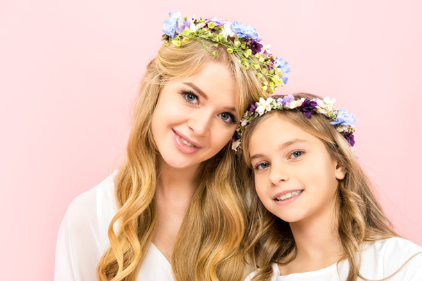 attractive mother and cute child in colorful floral wreaths looking at camera on pink background - Photo, image