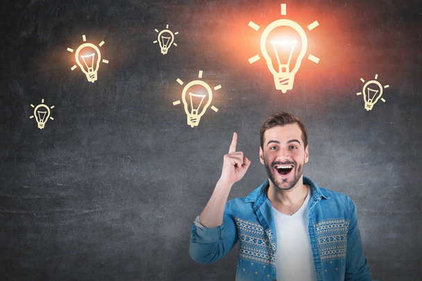 Portrait of happy young man in jeans shirt standing with open mouth and finger pointing up near blackboard with yellow light bulbs drawn on it. Concept of good idea - Φωτογραφία, εικόνα