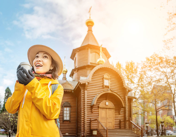 Happy tourist girl with hot drink travels in Russia and posing at the background of a old wooden church in authentic russian architecture - Photo, Image
