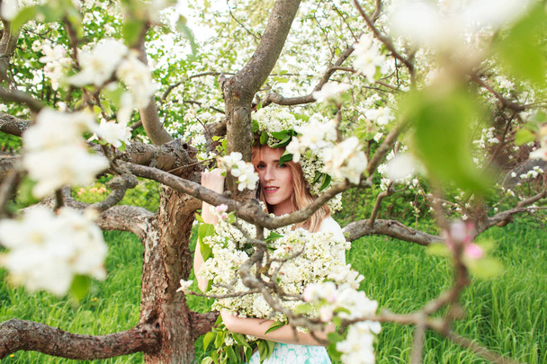 Young beautiful blonde woman in a dress in blooming apple garden outdoors. Many white blossom on the apple trees. The beauty of women, blossoming and spring. - Фото, изображение