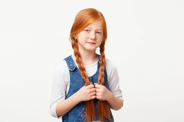 Tender little kind girl keeps in hands braided in two long plaits red hair, stands calmly directly looking camera, dressed in jeans overall dress over a white longsleeve isolated on white background - Fotografie, Obrázek
