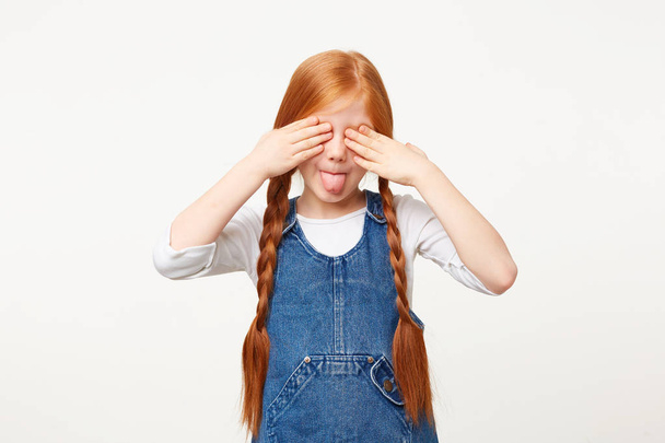 Mischievous funny little girl with red hair playing hide-and-seek with her parents standing closed eyes with palms on her eyes, shows tongue, dressed in a denim overall, over white background - Fotó, kép